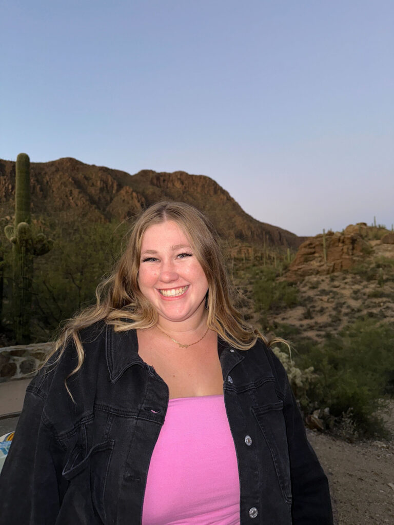 Vertical headshot of Emily against a Sonoran desert backdrop, smiling, wearing a pink shirt and black denim jacket